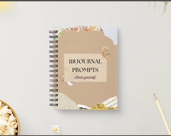 May include: A spiral-bound journal with a tan cover featuring the text "118 JOURNAL PROMPTS About yourself." The cover is decorated with torn paper and floral accents. A pencil and a bowl of popcorn are in the background.