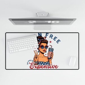 May include: A white desk mat with a retro illustration of a woman wearing sunglasses, a patriotic headband, and holding a drink. The mat has the text "N Free" and "But now I'm Expensive". A white keyboard and mouse are also visible.