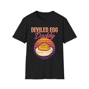 May include: Black t-shirt with a graphic of a deviled egg and the text "Deviled Egg Daddy" in a retro font. The design features a deviled egg on a sunset background with orange, yellow, purple, and pink stripes.