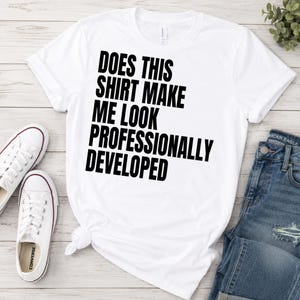 Op de afbeelding: Een wit t-shirt met korte mouwen en de zwarte tekst "DOES THIS SHIRT MAKE ME LOOK PROFESSIONALLY DEVELOPED" in een vet, schreefloos lettertype in het midden van de borst, gepresenteerd op een plat oppervlak.