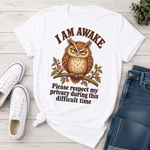 May include: A white short-sleeved t-shirt featuring a graphic of a grumpy brown owl perched on a branch with pink flowers. The text above the owl reads I AM AWAKE and the text below reads Please respect my privacy during this difficult time.