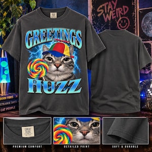 May include: A charcoal gray short-sleeve t-shirt featuring a graphic of a cat wearing a red, yellow, and blue propeller hat. The cat is next to a colorful swirl lollipop. The text GREETINGS and HUZZ appears in blue with a lightning background.