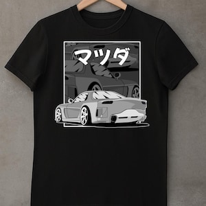 Op de afbeelding: Zwart t-shirt met een grijswaarden illustratie van een sportwagen en het Japanse woord "Mazda" in een witte rechthoek. Het ontwerp toont de achter- en zijkant van de auto tegen een vage achtergrond van vergelijkbare voertuigen.