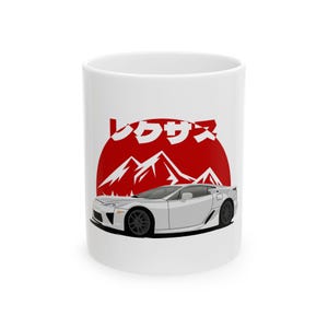 May include: White ceramic mug featuring a silver sports car design with black wheels. The car is set against a red sun and mountain graphic, with Japanese text above. A sleek, automotive-themed design.