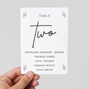 May include: A white table card with the word "Two" in elegant black script. The card also includes the word "TABLE" and a list of names. Decorative leaf illustrations are in each corner.