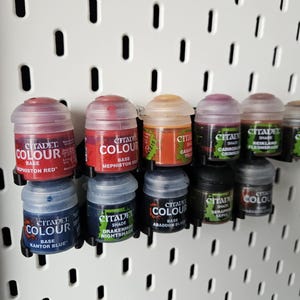 Puede incluir: Una colección de botes de pintura Citadel Colour en varios tonos, incluyendo rojo, azul y negro, exhibidos en una pared perforada blanca. Los botes están etiquetados y organizados para facilitar su uso.