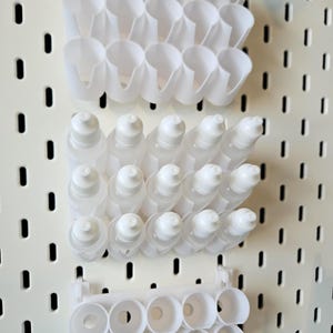 Peut inclure: Des organisateurs et des flacons en plastique blanc sont montés sur un panneau perforé blanc. Les organisateurs ont diverses formes, notamment des supports cylindriques et des compartiments incurvés. Les flacons ont des bouchons blancs et sont remplis d'un liquide clair.