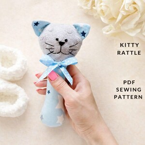 Könnte beinhalten: Ein graues Plüsch-Katzenrassel mit blauen Ohren mit Sternenmuster und einem passenden Griff. Die Rassel hat eine blaue Schleife und den Text "KITTY RATTLE PDF SEWING PATTERN".