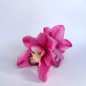 May include: A bright pink orchid flower with a light purple hair clip. The orchid has a white and brown centre with pink petals. The hair clip is a solid light purple colour and is positioned behind the flower.