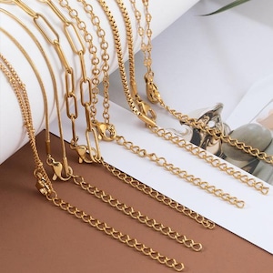 May include: A set of nine gold chain necklaces with different link styles and sizes. The chains are arranged in a fan shape on a light brown surface.