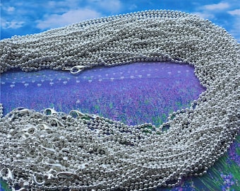 May include: A pile of silver ball chain necklaces with lobster clasps. The chains are arranged in a circular pattern on a purple and blue background.