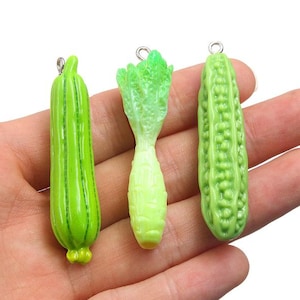 May include: Three miniature vegetable charms featuring a bright green cucumber with vertical ridges, a pale green leafy vegetable with a textured top, and a light green bitter melon with a bumpy surface, each with a small silver metal loop.