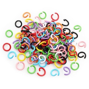 May include: A pile of colorful jump rings in various shades, including red, orange, yellow, green, blue, purple, pink, black, and white. The rings are made of a glossy material and are scattered across a white surface. These are ideal for jewelry making and crafts.