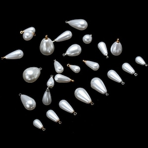 Peut inclure: Une collection de perles en forme de larme nacrées blanches avec des accents dorés et argentés. Les perles ont un trou en haut pour être enfilées ou attachées à des bijoux.