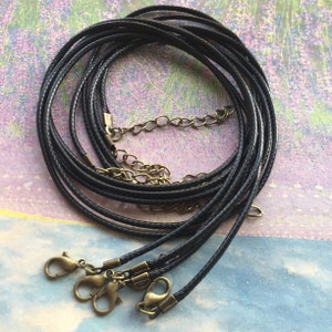 Peut inclure: Plusieurs colliers en cordon tressé noir avec des fermoirs et des chaînes d'extension de couleur laiton antique sont présentés. Les cordons sont bouclés et disposés sur un fond aux teintes violettes et bleues.