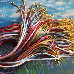May include: A large assortment of colorful satin cords with gold lobster clasps. The cords are arranged in a pile, with the clasps visible at the ends.