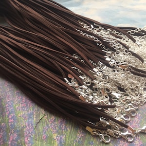 Pode incluir: Um conjunto de cordões de camurça castanho com fechos de lagosta prateados. Os cordões estão dispostos em uma pilha, com os fechos visíveis nas extremidades.