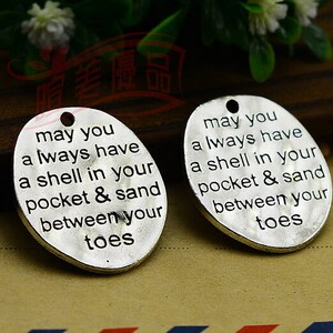 May include: Two oval-shaped, silver-colored pendants with a hole at the top. Each pendant has the engraved text: "may you always have a shell in your pocket & sand between your toes."