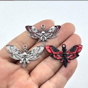 Puede incluir: Tres colgantes con forma de polilla con detalles de calavera. Dos son plateados, uno con detalles rojos. El tercero es negro con detalles de alas rojas. Cada uno tiene un lazo para colgar, ideal para joyería.