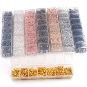 Op de afbeelding: Assortiment van kleine metalen sprongringen in verschillende kleuren en afwerkingen, georganiseerd in doorzichtige plastic containers. De containers zijn in een rij gerangschikt en zijn allemaal even groot. De sprongringen worden gebruikt voor het maken van sieraden en knutselen.