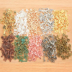 May include: A large collection of small metal key charms in various finishes including gold, silver, copper, bronze, green, and pink. The keys feature diverse decorative bow designs, intricate shafts, and varied bit shapes, arranged in piles.