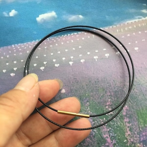 May include: A thin black cord necklace featuring a single cylindrical gold-toned metal bead connector. The necklace consists of two circular loops of black cord joined together by the metallic gold-colored tube component at the front center.
