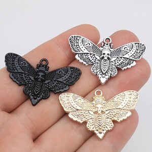 Puede incluir: Tres colgantes de metal con forma de polilla en negro, plateado y dorado. Cada colgante presenta un diseño de calavera en el cuerpo y intrincados patrones de alas. Los colgantes tienen un lazo en la parte superior para sujetarlos.