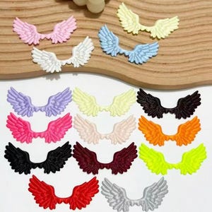 May include: A collection of small, flat, wing-shaped embellishments in various colours including pink, white, light blue, yellow, lavender, hot pink, cream, black, orange, burgundy, neon green, red, and silver, arranged in a scattered pattern.