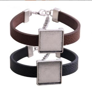 Könnte beinhalten: Zwei Lederarmbänder, eines braun und eines schwarz, mit quadratischen silberfarbenen Fassungen. Die Armbänder haben ein schlichtes Design mit einem Lederband und einem Metallverschluss. Die quadratischen Fassungen könnten personalisiert werden.