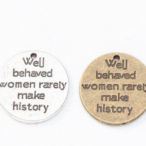 May include: Two round metal charms with the text "Well behaved women rarely make history" in a black font. One charm is silver and the other is gold.