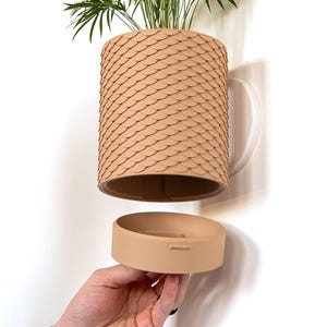 Peut inclure: Un pot de fleurs beige mural avec un motif en écailles. Le pot contient une plante verte et possède une soucoupe assortie. Le pot a une poignée blanche et est conçu pour être accroché au mur.