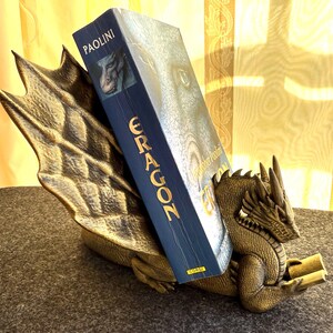 Op de afbeelding: Een bronzen draakvormige boekensteun met een boek getiteld "Eragon" van Christopher Paolini. De boekensteun heeft een gedetailleerd drakenontwerp met een getextureerde vleugel en een drakenkop. Het boek heeft een blauwe kaft.