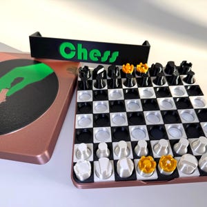 Op de afbeelding: Een draagbare schaakset met een koperkleurige koffer en een zwart-wit geblokt bord. De set bevat zwarte, witte en gouden schaakstukken. De koffer heeft een groene hand die een schaakstuk vasthoudt en het woord "Chess" in het groen.