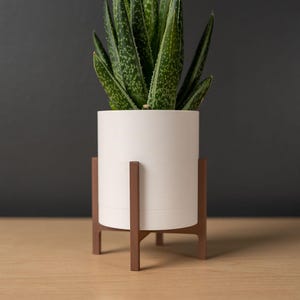 Op de afbeelding: Een witte cilindrische plantenbak met een groene plant, op een bruine houten standaard. De plant heeft groene bladeren met witte vlekken. De standaard heeft een modern design uit het midden van de eeuw.