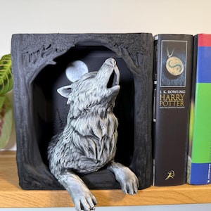 Könnte beinhalten: Eine graue Wolfsskulptur, die den Mond anheult, in einem dunklen, von Bäumen gesäumten Rahmen. Die Skulptur steht neben zwei "Harry Potter"-Büchern. Die Szene erinnert an ein mystisches Fantasy-Thema.