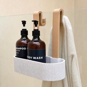 Op de afbeelding: Een lichtgrijze, ovale douchemand met een geribbelde textuur, voorzien van twee beige haken voor over de glaswand. In de mand staan twee amberkleurige flessen met zwarte pompdoseerders, gelabeld SHAMPOO en BODY WASH. Een beige handdoek hangt aan een haak.