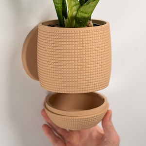 Op de afbeelding: Een bruinbeige, getextureerde keramische plantenpot met een bijpassende schotel en een aparte basis. De pot bevat een groene plant met lange, puntige bladeren. Het ontwerp heeft een rasterpatroon. De basis wordt door een hand vastgehouden.