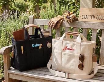 May include: A black canvas tote bag with the name Ushe embroidered in light blue and a cream canvas tote bag with the name Jessica embroidered in red. Both bags feature dual top handles, exterior pockets, and adjustable shoulder straps.