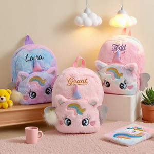 May include: Three plush unicorn backpacks in pink and blue hues, personalized with names. Each backpack features a unicorn face with a horn, wings, and a fluffy tail. A small pink mug and a unicorn-themed notebook are also present.