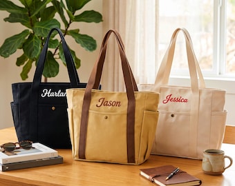 May include: Three canvas tote bags in black, tan, and cream, each featuring a front pocket with a snap button, side pockets, and two shoulder straps. The bags are personalized with embroidered names: Harlan in white, Jason in brown, and Jessica in red.