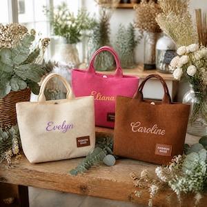 May include: Three corduroy tote bags in cream, hot pink, and brown. Each bag features two handles, a snap closure, and a rectangular brown patch labelled FASHION BAGS. The bags are embroidered with the names Evelyn, Eliana, and Caroline respectively.