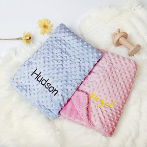Può includere: Due coperte per bambini, una azzurra e una rosa, con una superficie strutturata a pois. La coperta azzurra ha il nome "Hudson" ricamato in nero. La coperta rosa ha il nome "Hazel" ricamato in giallo. È visibile anche un giocattolo di legno.