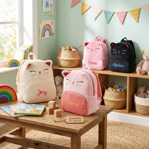 May include: Four plush cat-shaped backpacks in cream, light pink, and black, each featuring embroidered whiskers, ears, and a name or word like Joy, Love, Anne, or Madison. A wooden tag labeled HANDMADE KITTY BACKPACKS sits on the table.