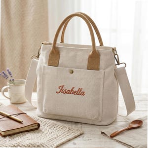 May include: A beige canvas tote bag with brown handles and a shoulder strap. The bag has a front pocket with the embroidered name "Tisabella" in red script. The bag is on a light-coloured wooden surface.
