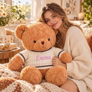 May include: A plush light brown teddy bear with a soft, textured fur finish. The bear is wearing a white knitted sweater featuring horizontal black and brown stripes at the collar and cuffs, with the name Emma embroidered in pink script text.