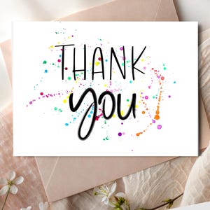 Puede incluir: Una tarjeta de felicitación blanca con las palabras "THANK YOU" en escritura negra y letras de bloque. La tarjeta presenta salpicaduras de pintura coloridas en tonos de azul, verde, rosa, naranja y amarillo. La tarjeta está sobre un sobre marrón claro.