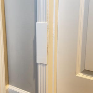 May include: A rectangular white plastic door frame filler piece attached to a vertical white door casing. The flat, smooth-surfaced component is positioned flush against the existing trim, extending the width of the molding to cover a gap.