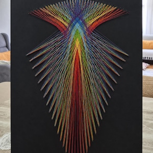 May include: A rectangular black board featuring a string art design composed of numerous thin, multicolored threads. The threads are arranged in a symmetrical, pointed pattern that radiates outward from the center, displaying a spectrum of colors.