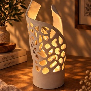 Peut inclure: Une lampe en céramique blanche avec un design ajouré composé de découpes irrégulières aux formes organiques. La structure cylindrique s'incurve vers le haut, laissant passer une lumière chaude à travers les ouvertures.