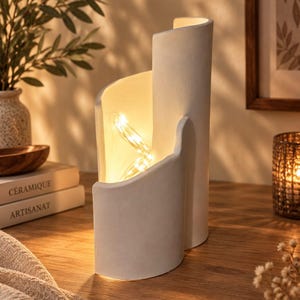 Peut inclure: Une lampe sculpturale en céramique blanc mat avec une silhouette incurvée à plusieurs niveaux. L'intérieur de la structure est éclairé par une guirlande de petites lumières chaudes qui projettent une lueur douce sur les parois incurvées.
